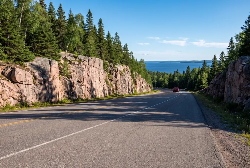 Illustration: a two-lane highway curving between pink granite rock cuts and spruce forest, with a big blue lake beyond the trees.